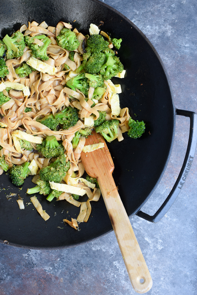 Stir-fried shirataki noodles with colorful vegetables and garnishes, served in a shallow white bowl with chopsticks resting on the edge.