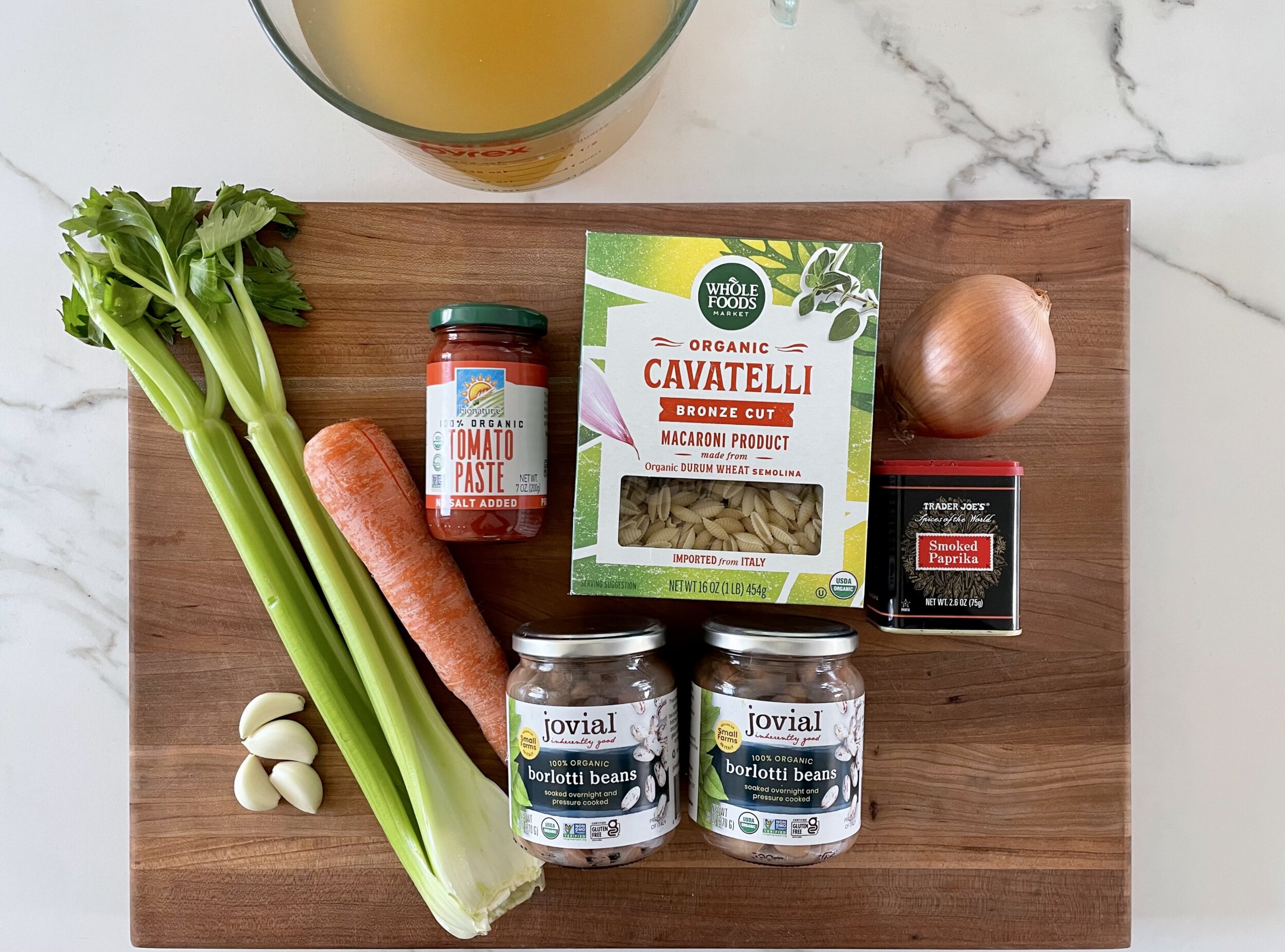 Ingredients for pasta e fagioli soup displayed on a wooden cutting board placed on a white surface, including celery, carrots, onion, garlic, white beans, tomato paste, and cavatelli pasta.