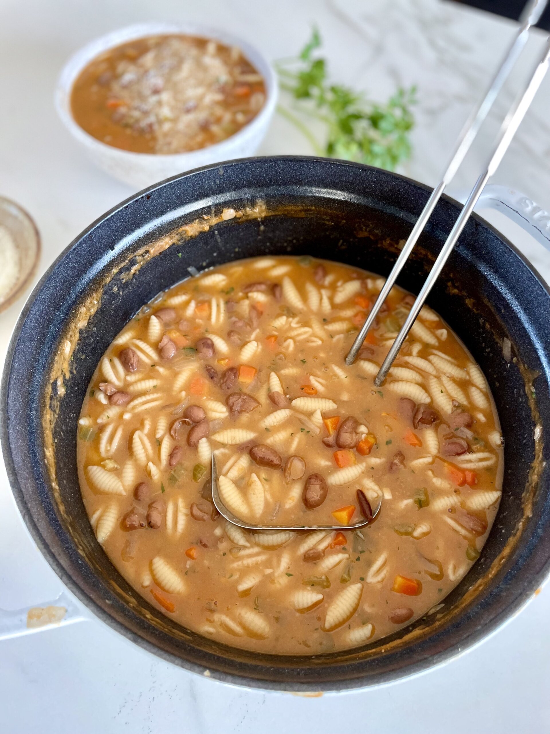 Hearty pasta e fagioli soup in a large pot with a ladle in it filled with beans, pasta, and vegetables in a rich tomato broth. Behind the pot is a white bowl filled with pasta e fagioli soup.