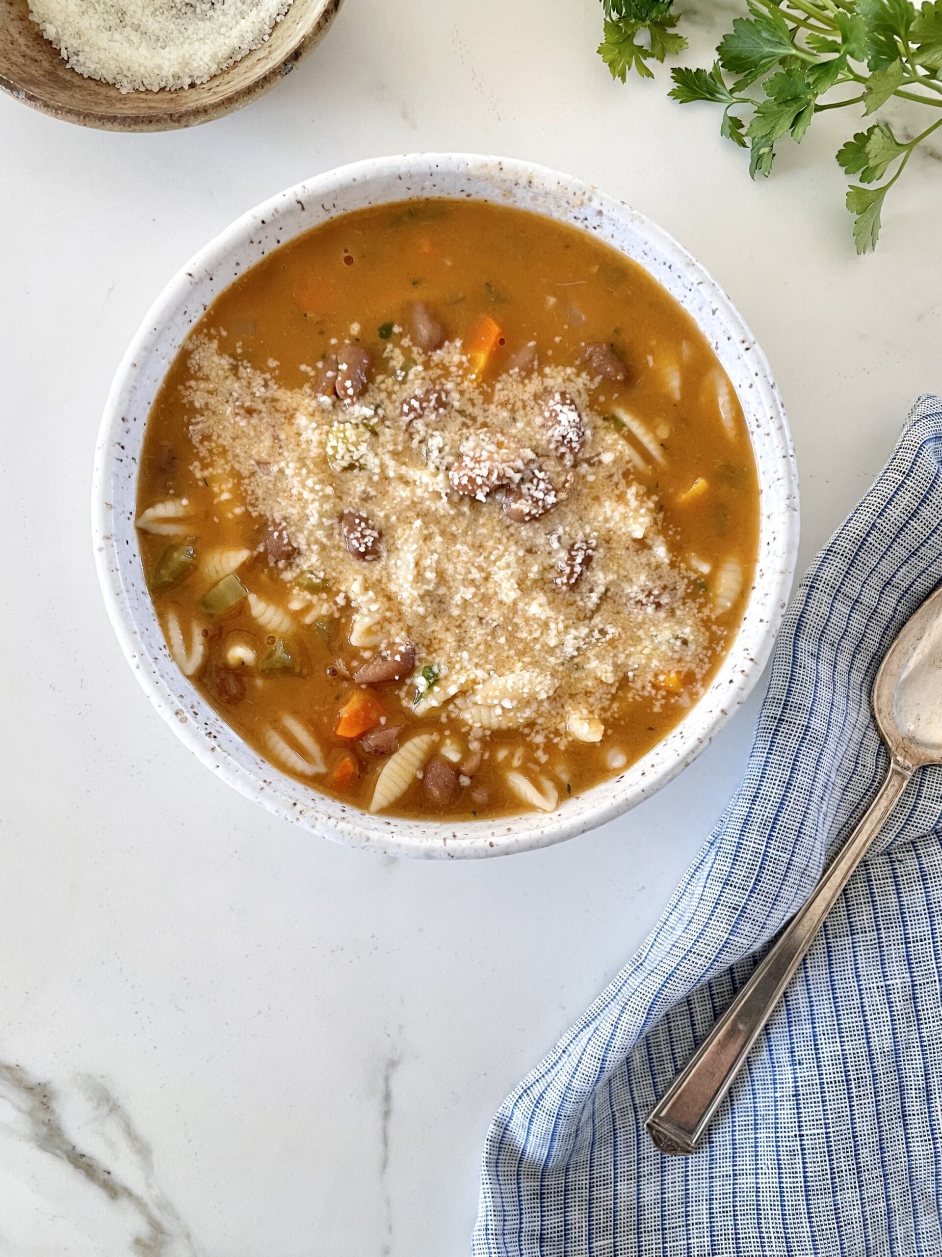 Close-up of pasta e fagioli soup with tender pasta, creamy beans, and chunky vegetables in a deep, savory broth. A blue kitchen towel and a silver spoon sit next to the bowl.