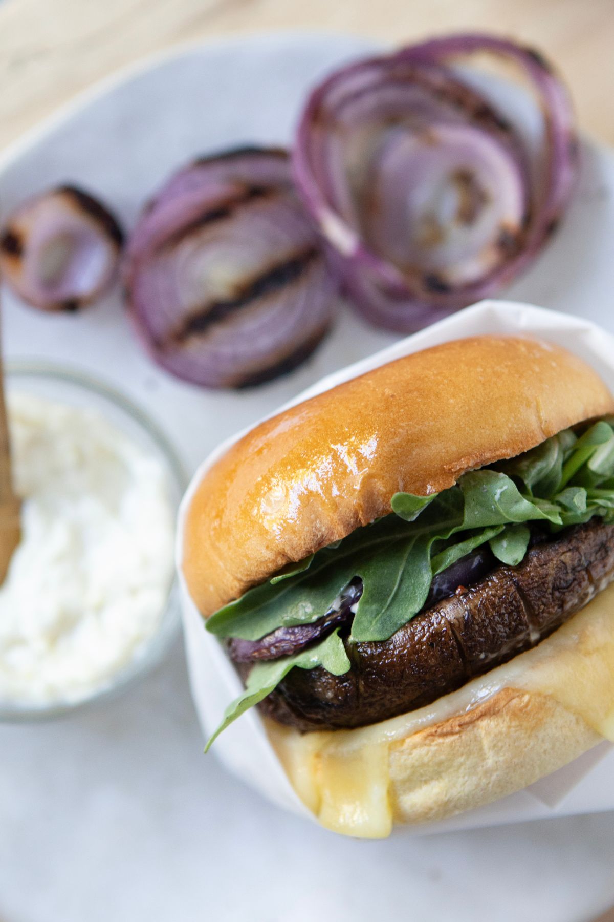 Assembled grilled portobello burger with aioli, cheese, grilled onion, and arugula on a toasted bun.