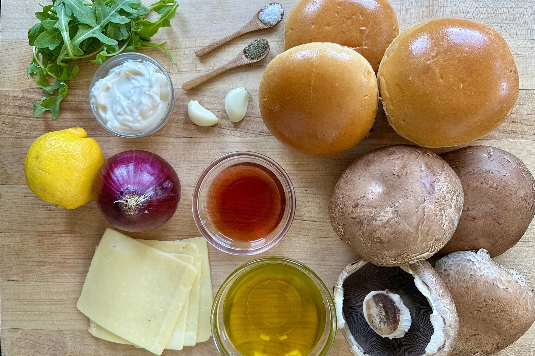 Ingredients for grilled portobello burgers including portobello caps, arugula, red onion, garlic-chive aioli, and buns laid out on a prep board.