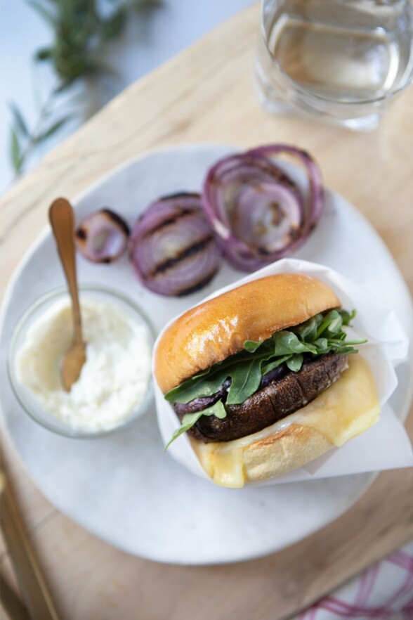 Assembled grilled portobello burger with aioli, cheese, grilled onion, and arugula on a toasted bun.

