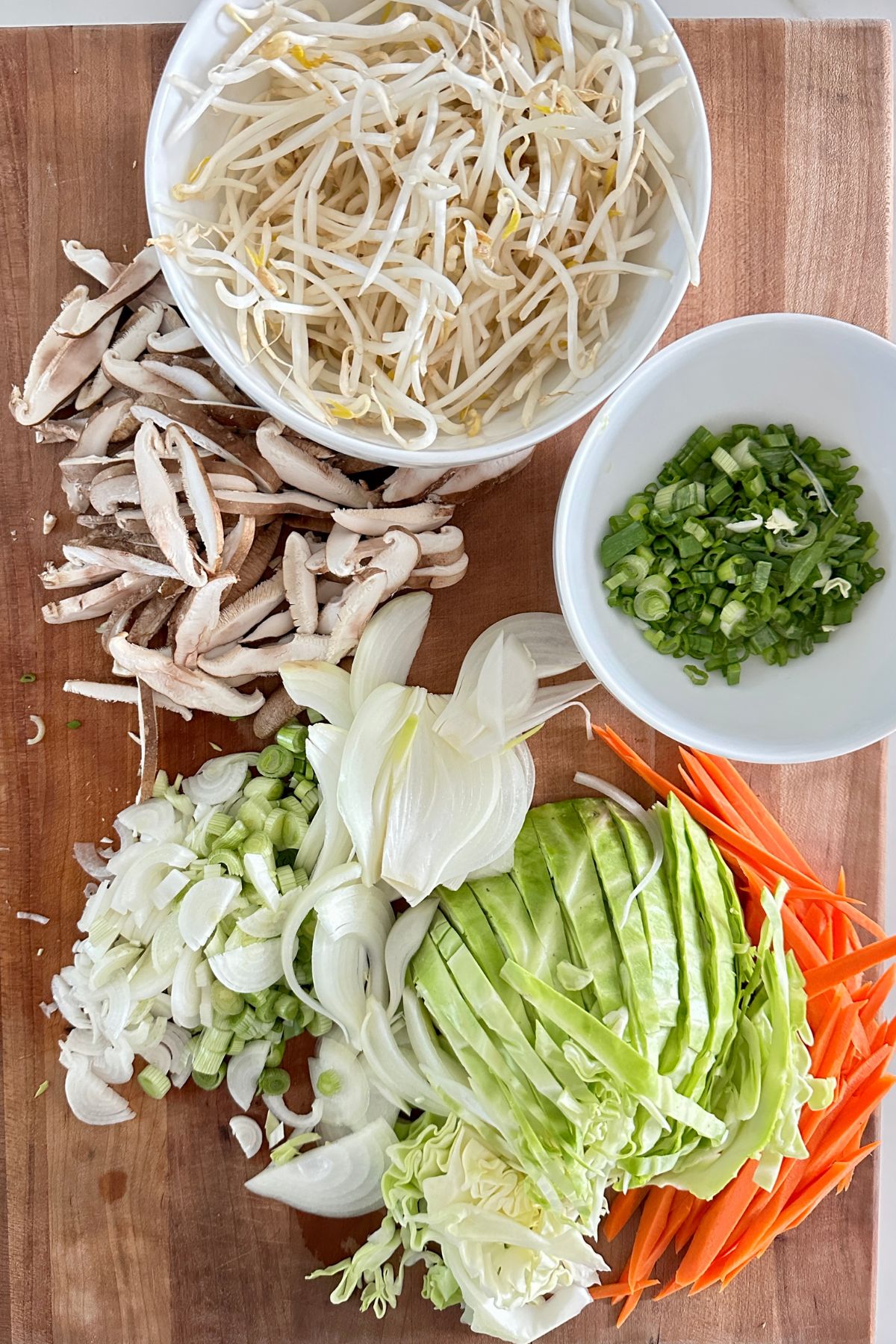 Sliced vegetables for shrimp lo mein, including cabbage, scallions, mushrooms, and carrots, arranged on a cutting board