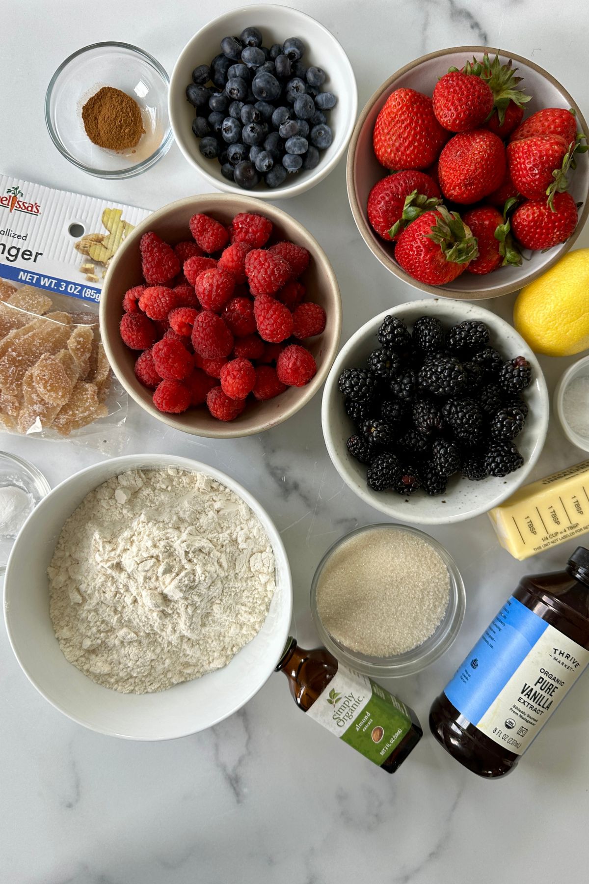 Overhead view of small bowls and ramekins holding unbleached and whole wheat flours, cane sugar, sea salt, baking powder, ground cinnamon, melted butter, cold water, fresh mixed berries, lemon, arrowroot starch, vanilla extract, and almond extract arranged for a mixed berry crumble pie
