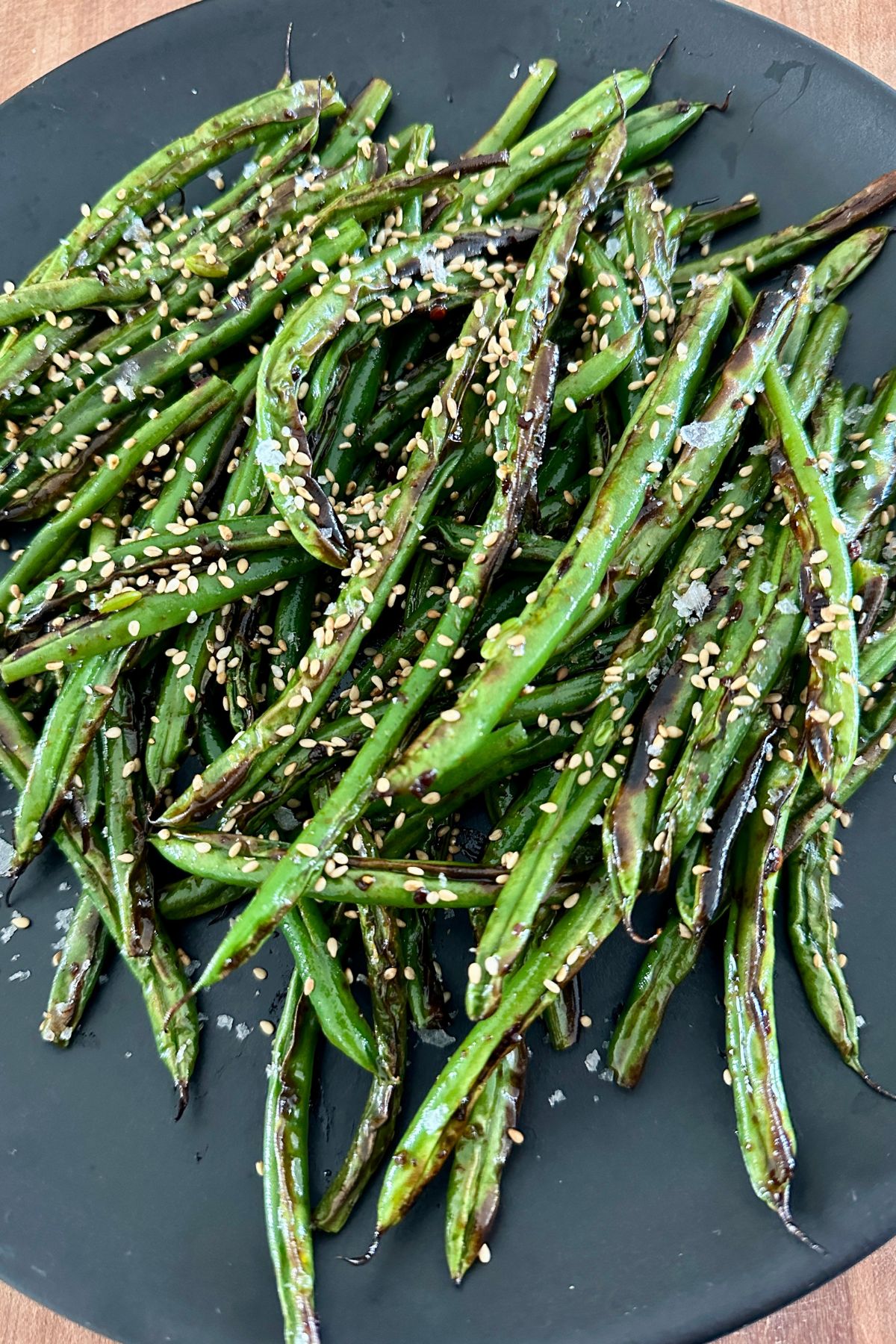 a close up of blistered green beans cooked and seasoned on a black plate and garnished with sesame seeds