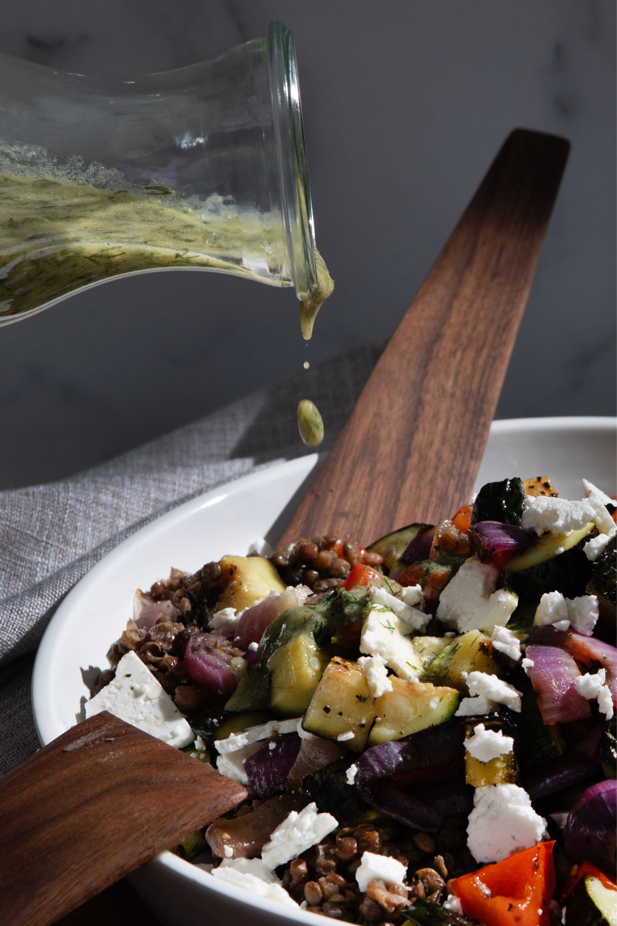 Lemon dill dressing being poured over a grilled veggie salad in a white bowl with a dark wooden spoon with a dark background.