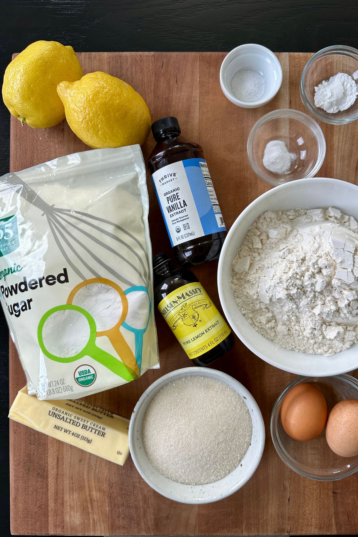 ingredients to make lemon crinkle cookies on a cutting board including fresh lemons, all purpose flour, sugar, and eggs
