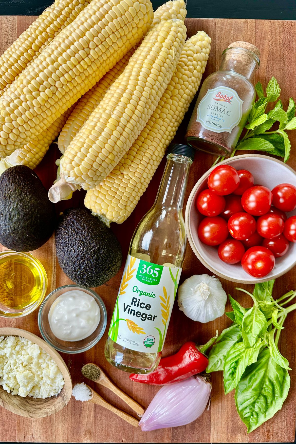 Ingredients for summer corn salad laid on a dark wooden cutting board including ears of corn, avocado, cherry tomatoes, and seasonings. 