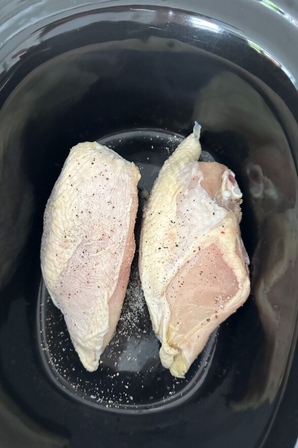 Seasoned raw chicken breasts laying in a black crock pot ready to cook.
