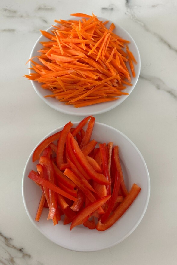 Shredded carrots and sliced red bell peppers in two small white bowls, set against a clean white background.