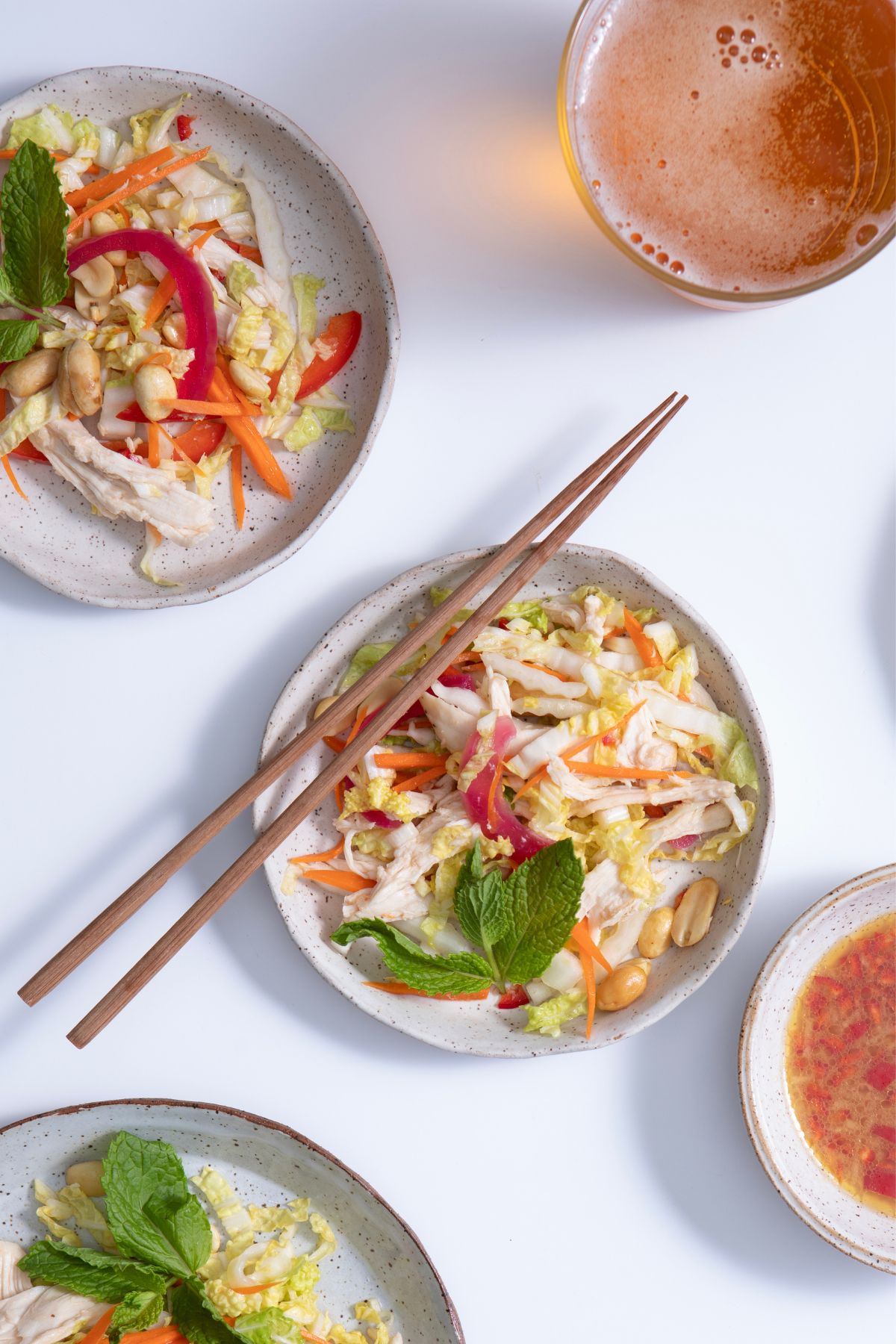 Three shallow speckled white bowls filled with Vietnamese chicken salad, garnished with fresh herbs. Chopsticks rest on the main bowl, all set against a clean white background