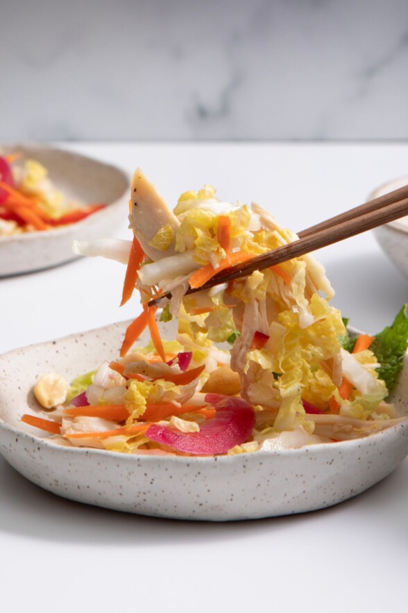 Vietnamese chicken salad in a speckled white bowl, being picked up with chopsticks against a soft, light-toned white and gray background.